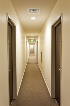 Long empty corridor in a hotel or apartment building interior with overhead lighting, doors on either side and illuminated exit sign