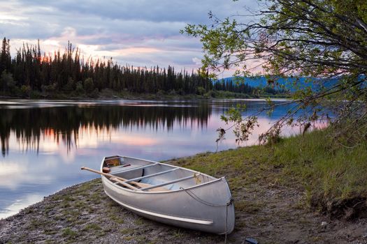 Canoe and paddles beached on shore of beautiful Teslin River in the remote wilderness of Yukon Territory, Canada, the river surface reflecting delicate sunset colors