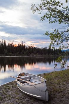 Canoe and paddles beached on shore of beautiful Teslin River in the remote wilderness of Yukon Territory, Canada, the river surface reflecting delicate sunset colors