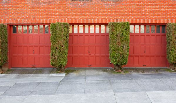 Three Red Garages on a brick wall separaged by evergreen bushes