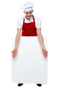 Handsome young cook showing blank billboard against white. Cheerful cook