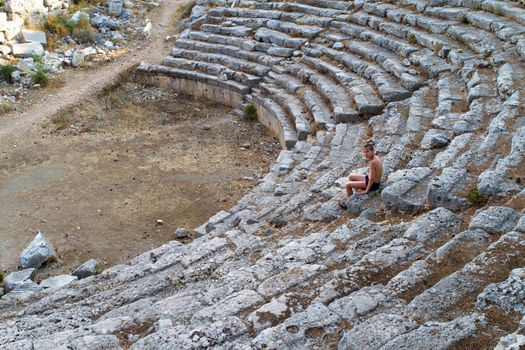 Boy sitting on steps of stone Ancient amphitheater. Phaselis, Turkey
