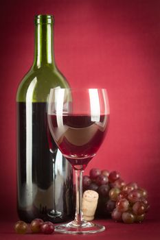 A bottle of red wine, a glass half full and grapes on a vintage red background.