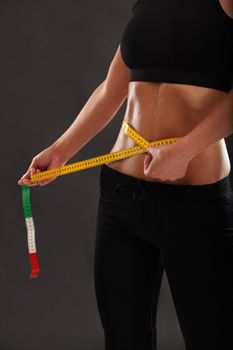 Photo of a tanned slim young woman measuring her sweaty waistline over dark background.