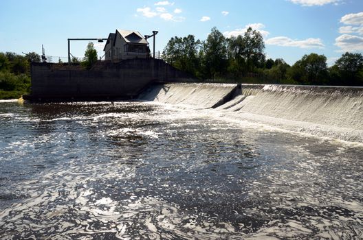 Dirty barmy prothy river water flow through weir dam barrage. House on shore.