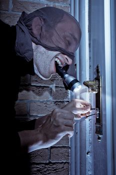 Masked burglar holding a flashlight in his mouth, picking the lock of a door.