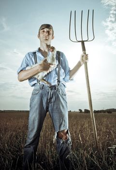 Young farmer standing in farm field with chicken and a pitchfork.