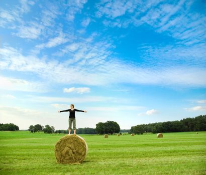woman balancing on top of a hay bale on a meadow, infront of  a blue sky