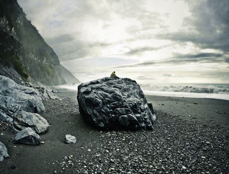 Man sitting on a big rock on a rocky beach watching the ocean. Location: Hualien County, Taiwan