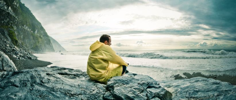 Young man sitting on a rock on a rocky beach watching the ocean in a yellow rain coat. Location: Hualien County, Taiwan