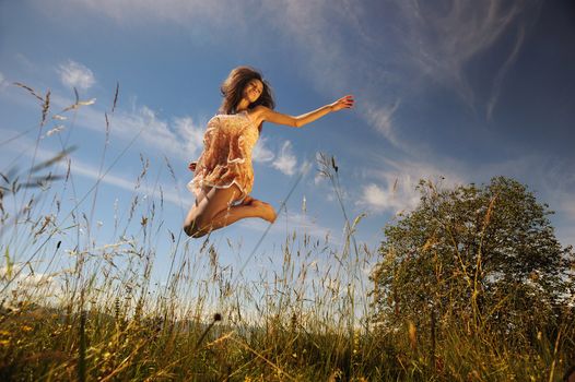 a smiling young woman in a green lawn makes a jump with open arms, she is silhouetted against the blue sky