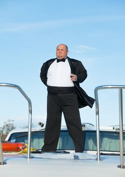 Overweight man in tuxedo standing on the deck of a luxury pleasure boat with glass red wine