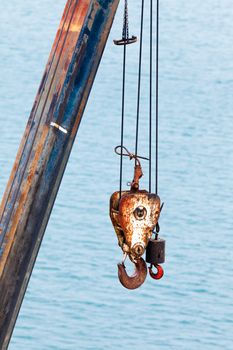 Two different sized battered and rusted hooks and cables of crane for hoisting or lifting heavy loads in harbor against blurry ocean background