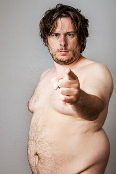 An overweight man who is naked pointing at the camera firmly. Man is shabby looking with unkempt hair and a rough beard. 