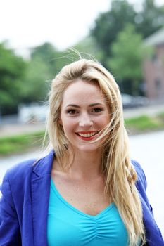 Beautiful vivacious young blonde woman with a lovely smile standing outdoors in a blue top and jacket 