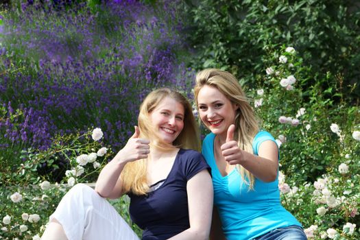 Happy motivated friends sitting close together amongst garden flowers giving thumbs up of approval and hope 