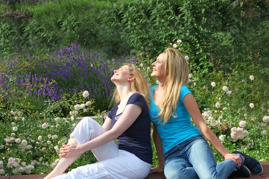 Two pretty young women sitting close together in a lush garden with flowers looking up at the sky and enjoying the sunshine 