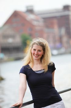 Friendly smiling blonde woman leaning against a railing outdoors above a river with the breeze in her hair 