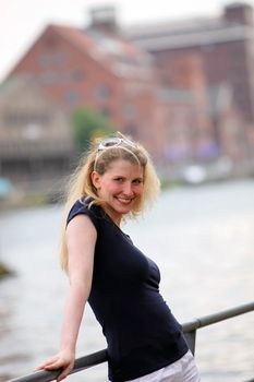 Smiling attractive blonde woman in a casual black top leaning against a railing overlooking a river with copyspace 