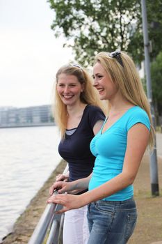 Two smiling attractive female friends standing talking at a railing alongside a river Two smiling attractive female friends standing talking at a railing alongside a river