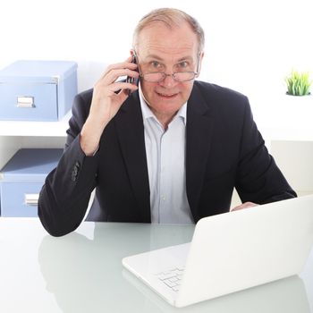 Hardworking middle-aged businessman sitting at his desk in front of his laptop talking on his mobile phone 