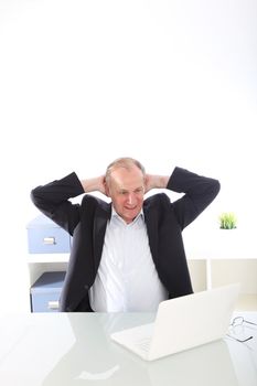 Satisfied businessman relaxing back in his chair with his arms raised behind his head as he looks at his laptop 