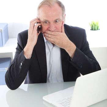Businessman seated at his desk covering his mouth with his hand in shock as he receives bad news over his mobile phone 