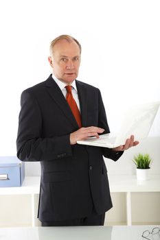 Senior stylish business executive standing working on his laptop which is balanced on his arm as he waits for the start of a meeting 