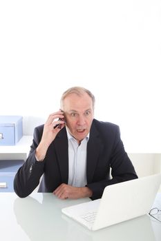 Middle-aged businessman seated at his desk reacting to news received over his mobile phone in disbelief 