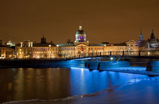 Night time view of Bonsecures Market in Montreal, Quebec, Canada, North America. Photo shows the entire facade of the landmark building. View is from across the river showing the foot bridge and blue light reflecting from the water. Season is winter.
