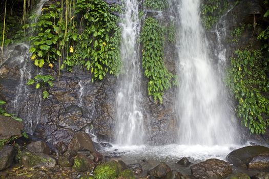 Twin waterfall cascading down a rocky cliffside covered with tropical, green vegetation.