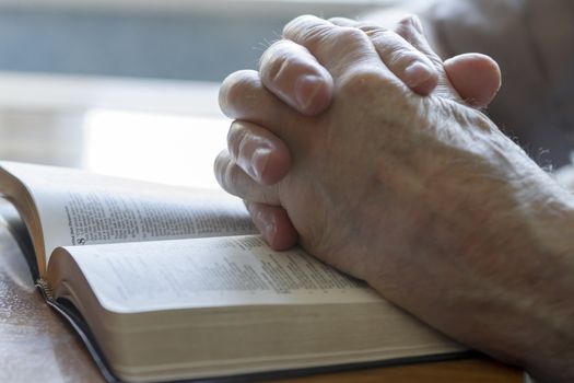 weathered old man's hands clasped in prayer over open Bible