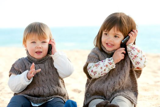 Portrait of two cute little girls on beach having conversation on mobile phones.