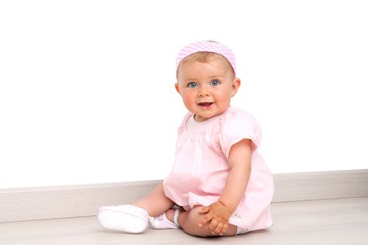 Friendly blue eyed baby girl wearing pink dress and pink head band sitting on the floor indoors