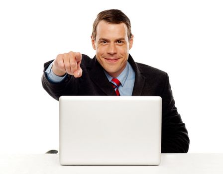 Handsome male executive pointing at you while working on laptop at his work desk