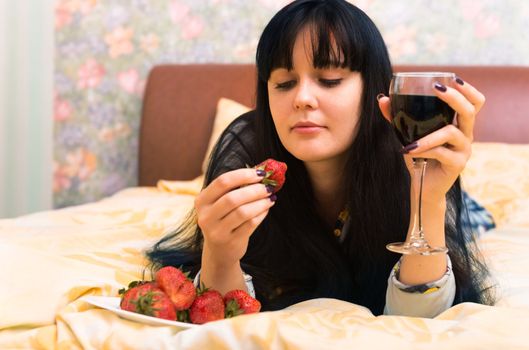 Happy young woman in bed with strawberries and glass of wine