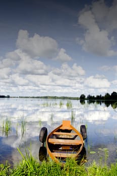 Boat moored to shore between rubber tires. Amazing summer lake view and cloudy sky reflections on water.