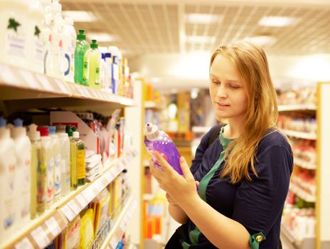 Young blonde woman is reading inscription on the cleaner bottle in the supermarket