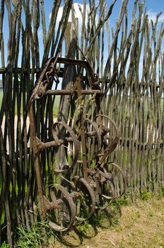 Old corroded harrowing field the tool stand near handmade fence from tree branches.