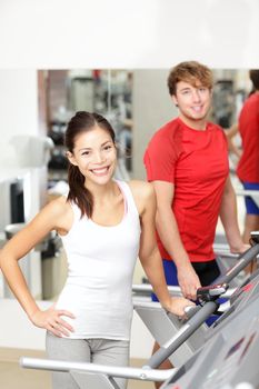 Fitness people. Couple running on treadmill indoors in gym fitness center. Young woman fitness model smiling with young man behind.