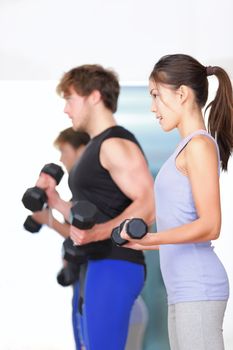 Fitness people in gym. Couple strength training lifting weights during indoor fitness workout. Woman lifting dumbbells training biceps in focus.