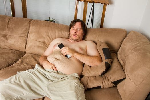 Fat obese man asleep on the couch with remote in one hand and the other hand shoved down the front of his pants. Shows obesity due to lack of excercise.
