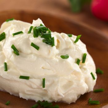 Fresh cream cheese spread on wooden plate with chives on top and radish in the back (Selective Focus, Focus on the chives on the top of the cream cheese)