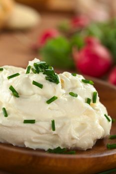 Fresh cream cheese spread on wooden plate with chives on top and radish in the back (Selective Focus, Focus on the chives on the top of the cream cheese)