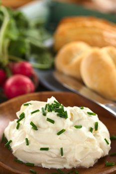 Fresh cream cheese spread on wooden plate with chives on top, radish and buns in the back (Selective Focus, Focus on the chives on the top of the cream cheese)