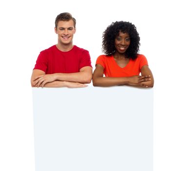 Smiling young couple standing behind white billboard looking at camera