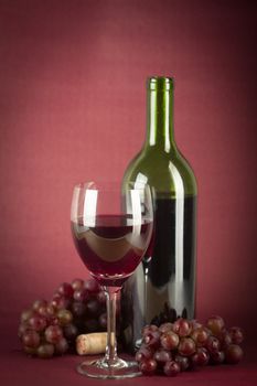 A bottle of red wine, a glass half full and grapes on a vintage red background.