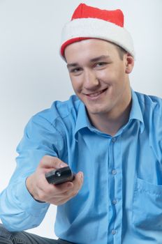 The young and handsome man in a red cap and a festive blue shirt on a white background, watching Christmas TV holding a TV remote