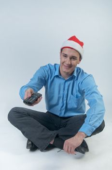 The young and handsome man in a red cap and a festive blue shirt on a white background, watching Christmas TV holding a TV remote