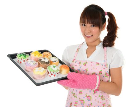 Young Asian girl baking bread and cupcakes, wearing apron and gloves holding tray isolated on white.